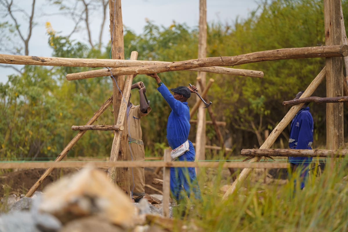 Workers in blue uniforms construct the wooden framework of a building at the Kibale Lodge, contributing to the sustainable development of Volcanoes Safaris' newest eco-lodge in Uganda.