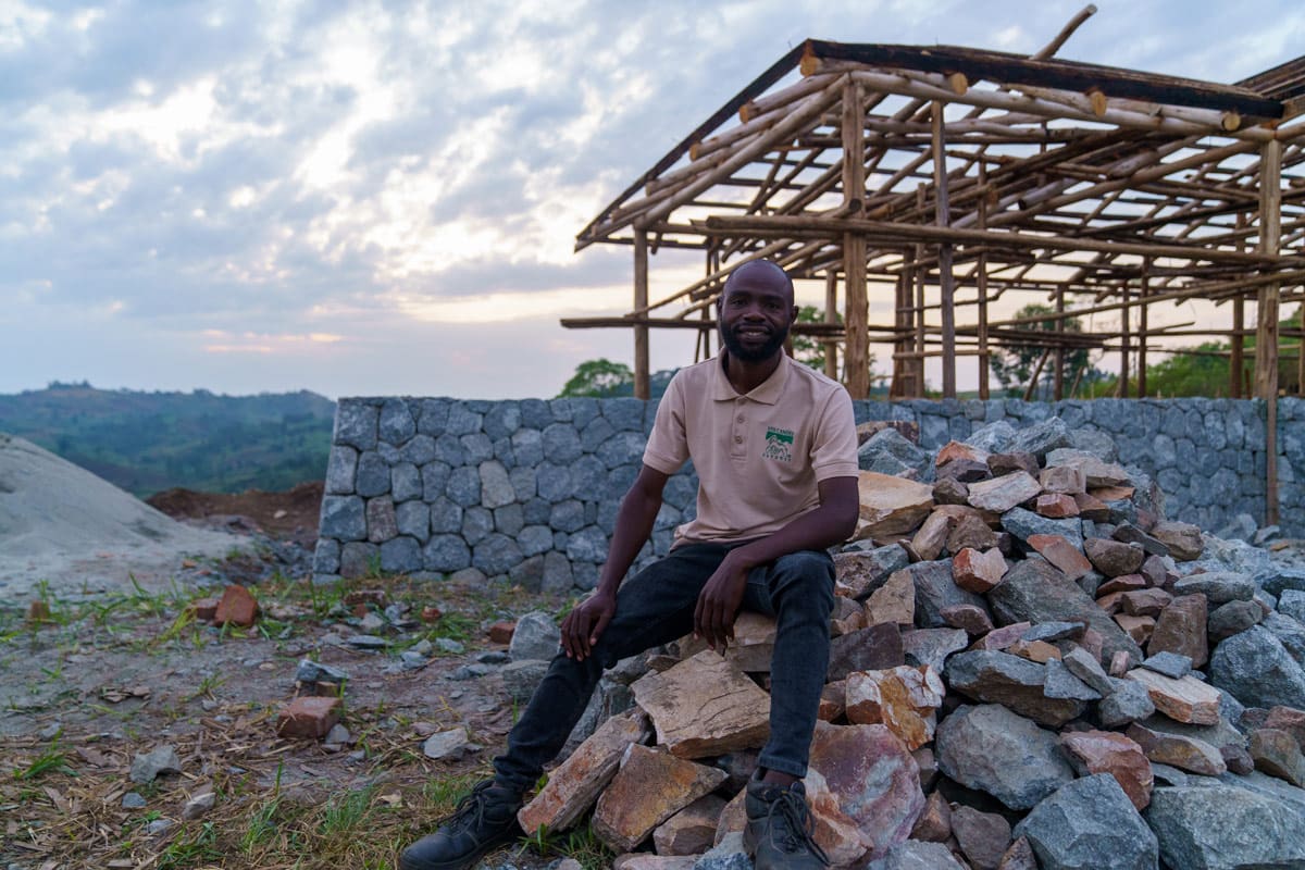 JB, the construction supervisor at Volcanoes Safaris, takes a moment to rest on a pile of stones with the wooden framework of the Kibale Lodge rising in the background, as the day fades in Uganda.